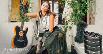Woman holding guitar and wearing luxury brand clothing from the Thale Blanc trunk show. Designer shoulder ba in black leather with metal studs sits nearby.
